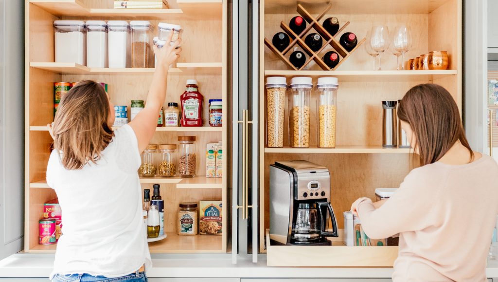 Image Description: Kitt and Joni stand with their backs facing us, while they organize open pantry cabinets