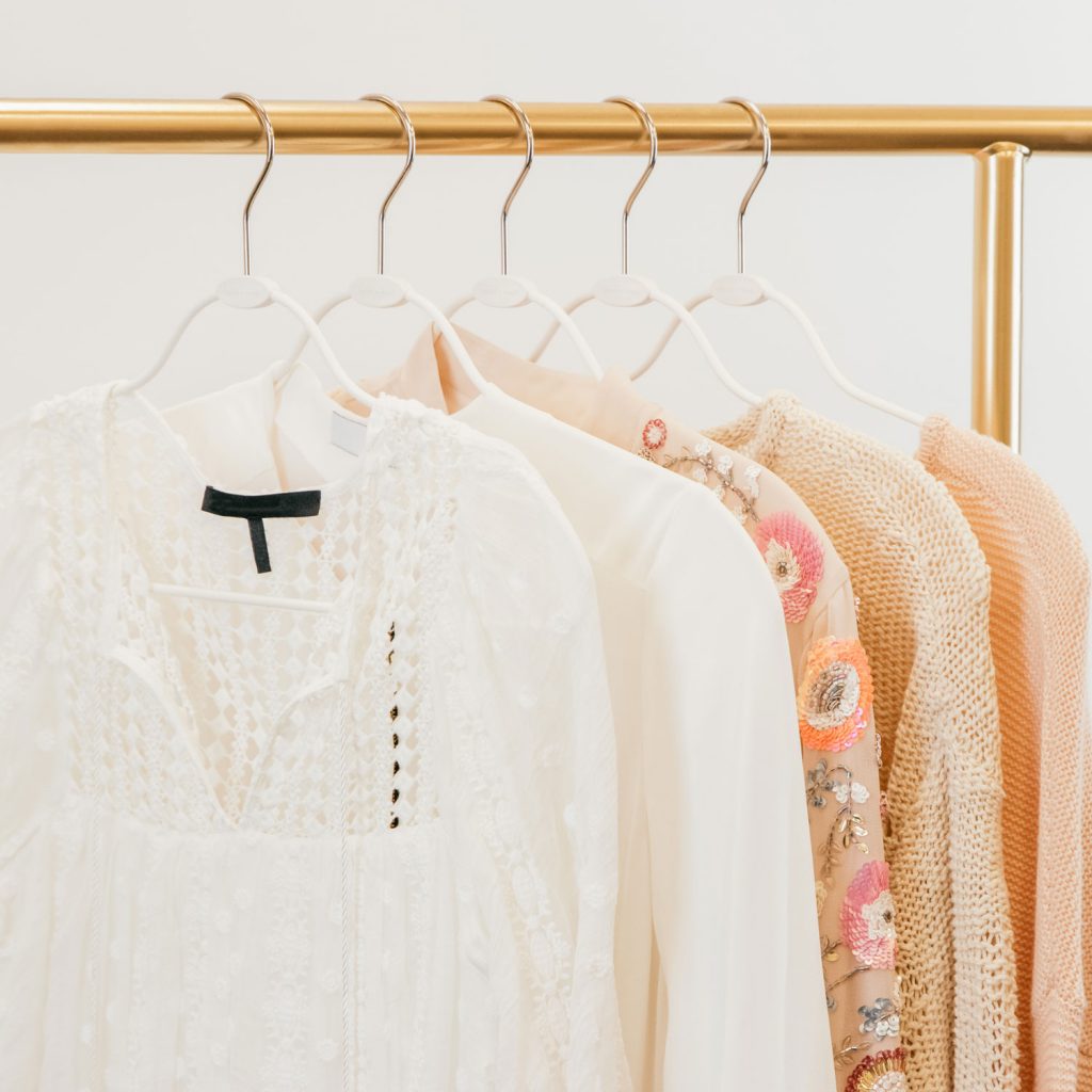 Image Description: Slim white hangers are showcased on a gold clothing rod, with light colored blouses