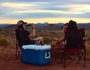 Image Description: Two friends sit in camping chairs in the desert with a blue Coleman cooler between them