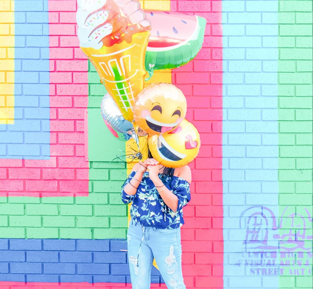 Image Description: A woman holding happy face balloons in front of her face stands in front of a multi-colored wall