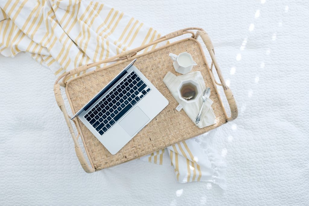 Image Description: A laptop and cup of tea rest on a wicker tray, atop a white bedspread
